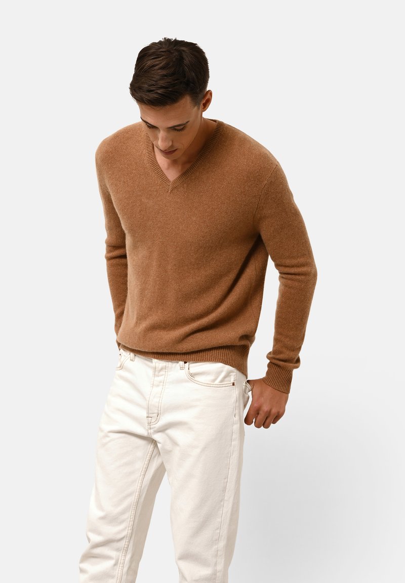 V-Ausschnitt Pullover aus leichtem braunem Wollmischung, mit gerippten Ärmelbündchen und Saum, kombiniert mit weißen Jeans. Glatte Textur mit einer figurbetonten Silhouette.