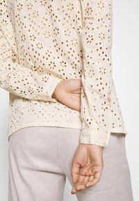 Blusa ricamata color beige chiaro con ritagli floreali, maniche lunghe e orlo arrotondato, abbinata a pantaloni di colore chiaro.
