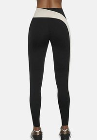 Widok z tyłu osoby w czarnych legginsach z beżowym ukośnym pasem owiniętym wokół talii i nogi, połączonych ze metalicznie złotymi butami sportowymi.