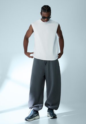 Mann mit geflochtenem Haar, der eine Sonnenbrille, ein ärmelloses weißes Hemd, lockere dunkelgraue Hosen und Turnschuhe trägt und den Saum seines Hemdes nach unten blickend richtet.