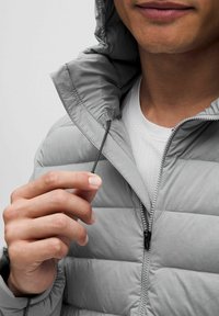 Svetlo siva puhovka z zadrgo na sprednji strani in visokim ovratnikom. Tkanina je gladka z rahlo sijočim finišem. Opazna je tudi nastavljiva vrvica.