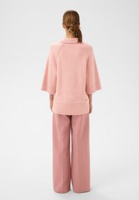 Žena s hnědými vlasy svázanými do culíku, oblečená v volném růžovém svetru a širokých růžových kalhotách, stojí zády k objektivu na jednobarevném pozadí.