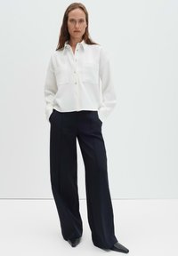 Camicia bianca con bottoni in tessuto strutturato, tasche sul petto; abbinata a pantaloni neri a gamba larga e scarpe nere a punta.