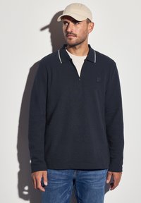 Tmavě navy modrá polokošile s dlouhými rukávy a předním zipem do půlky, kontrastním šedým lemem límce a malým logem na hrudi, spárovaná se světle modrými džíny a béžovou kšiltovkou.