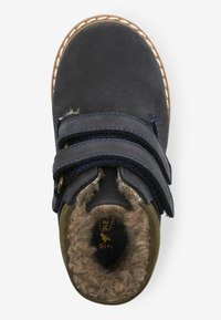 Mørkeblå ruskindsstøvle med to sorte velcro-stropper, pelsforing og syet gummisål. Runde tå og olivengrønne detaljer på siden.