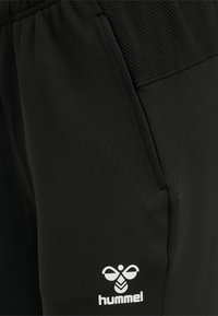 Schwarze Sporthosen aus Stretchstoff mit einem Netzbund und einem kleinen weißen Logo auf der unteren linken Seite.