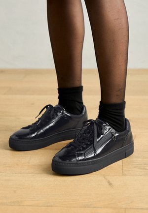 Schwarze Lackledersneaker mit Schnürverschluss, strukturiertem Obermaterial und gepolsterter Sohle. Getragen mit schwarzen Knöchelsocken. Flaches, rundes Zehendesign.