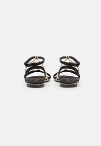 Black strappy sandals featuring a flat heel, textured suede material, gold accents, and a buckled ankle strap for secure fit.