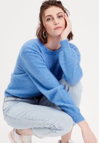 Pull bleu clair en matériau doux, avec des manches longues et un col rond, associé à un jean en denim clair et des baskets.