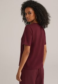 Weinrote, strukturierte Bluse mit kurzen Ärmeln und rundem Halsausschnitt, getragen mit passenden Hosen. Einfaches Design, sichtbare Nähte und lässiger Schnitt.
