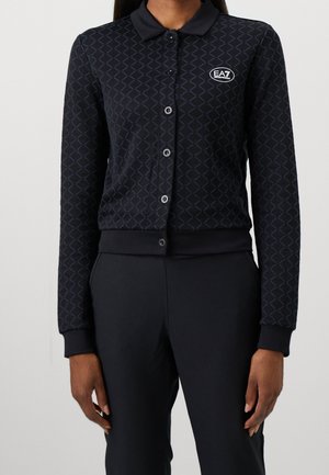 Femme portant un haut noir à manches longues boutonné avec un motif subtil en losange et le logo EA7 sur la poitrine, associé à un pantalon noir.