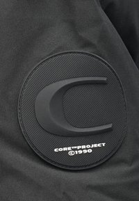 Mustast tekstiilist kangas, millel on ümmargune, eest kõrgendatud kummist plaastri tekstuurse diagonaalse mustri ja reljeefse logotekstiga "CORE™ PROJECT ©1990".