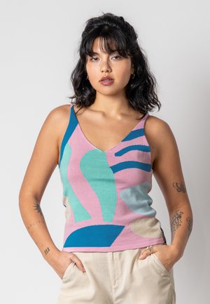 Woman with dark wavy hair wearing a colorful abstract-patterned tank top and light pants, standing with hands in pockets against a plain background.
