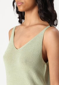 Lys grønn, ermeløs strikketopp med V-hals og en myk, teksturert veving. Subtil glans gir stoffet visuell interesse.