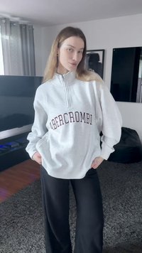 Svetlo siva flis jopa z ovratnikom na polovični zadrgo, z temnordečim logotipom spredaj. Kombinirana s črnimi širokimi hlačami.