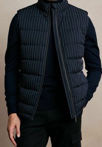 Chaleco acolchado azul marino con rayas blancas verticales, cuello alto, cierre de cremallera frontal y un diseño elegante, combinado con un suéter oscuro debajo.