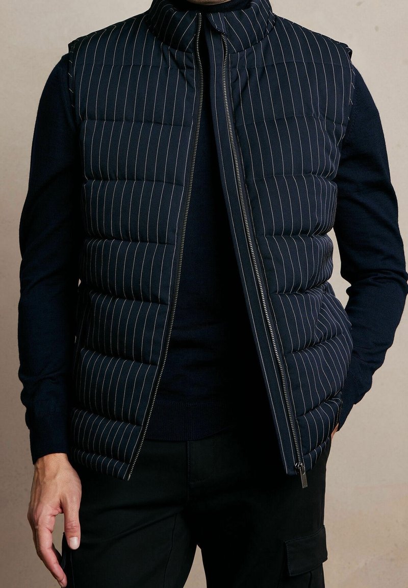 Chaleco acolchado azul marino con rayas blancas verticales, cuello alto, cierre de cremallera frontal y un diseño elegante, combinado con un suéter oscuro debajo.