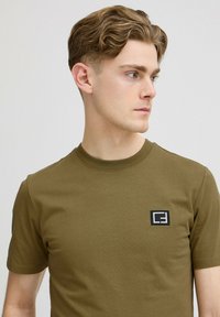 Tricou din bumbac de culoare verde măsliniu, cu guler rotund, mâneci scurte și un mic patch dreptunghiular negru cu logo pe pieptul stâng.