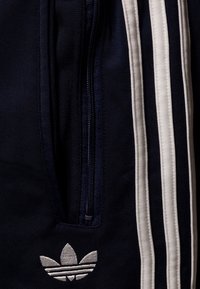 Navy blue athletic pants with white stripes, featuring a zippered pocket and a silver embroidered logo at the lower hem.
