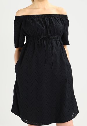 Schwarzes schulterfreies Kleid mit kurzen Ärmeln, tailliert, mit Taschen und Lochmuster, getragen von einer Person vor einem einfarbigen Hintergrund.