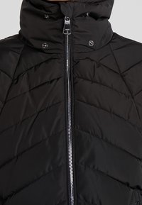 Veste matelassée noire avec couture en chevrons, fermeture éclair argentée à l'avant, col montant et fermetures à boutons-pression près du cou.