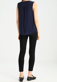 En person står med ryggen mot betraktaren, iklädd en ärmlös marinblå topp, svarta skinny jeans och svarta ballerinas framför en enhetlig bakgrund.