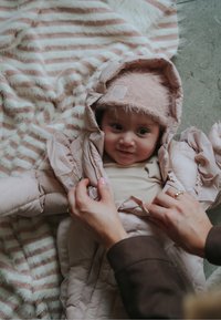 Vaaleanpunainen toppatakki hupulla, pehmeästä kankaasta ja vetoketjulla suljettava, päällä beige paita. Taustalla raidallinen viltti.