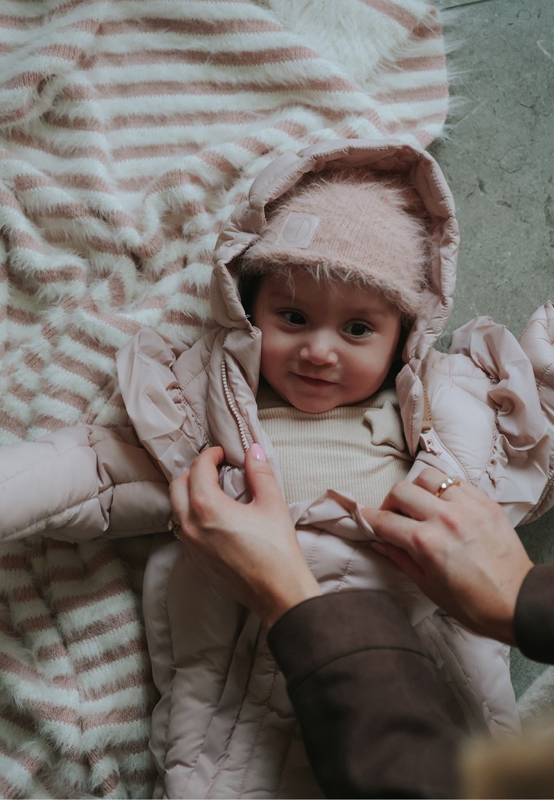 Vaaleanpunainen toppatakki hupulla, pehmeästä kankaasta ja vetoketjulla suljettava, päällä beige paita. Taustalla raidallinen viltti.