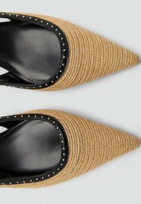 Buty z szpiczastym noskiem wykonane z plecionego beżowego materiału, z czarnymi skórzanymi wykończeniami oraz małymi metalowymi ćwiekami wzdłuż krawędzi, charakteryzujące się teksturowaną powierzchnią.