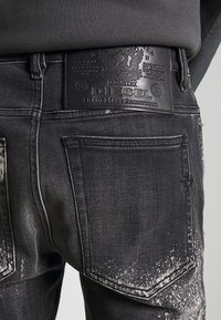 Vaqueros de mezclilla negra con un patrón desgastado y rasgado, que presentan un parche de cuero de la marca Diesel en la cintura, con un estilo de bolsillo tradicional.