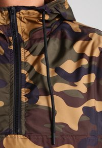 Cazadora de pullover en camouflage de color verde, marrón y beige. Cuenta con un alto cuello, cremallera negra y cordón ajustable en el cuello.