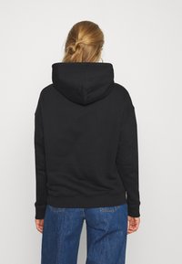 Sudadera negra con capucha y corte relajado, con puños y dobladillo acanalados, combinada con vaqueros de pierna ancha en azul. La tela parece suave y texturizada.