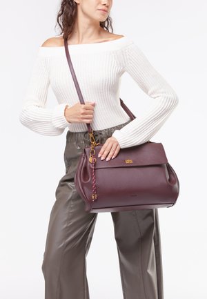 Borsa in pelle borgogna con un design strutturato, dettagli in hardware dorato e una tracolla intrecciata. Modello indossa un maglione bianco a coste.