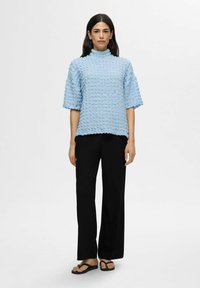 Lyseblå tekstureret bluse med høj hals og cropped ærmer, parret med sorte breddebenede bukser og sorte klipklappere.