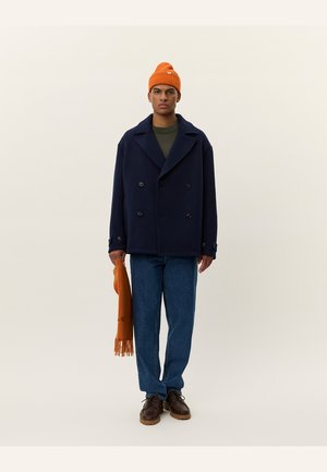 Navy double-breasted peacoat over a green sweater, blue jeans, brown shoes, and an orange knit beanie with a scarf in hand.