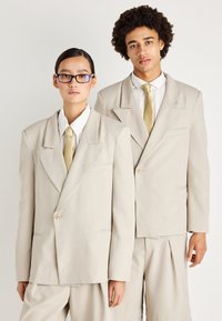 Dois modelos vestem fatos oversized em bege com lapelas marcadas, botões à frente e gravatas amarelo pálido a condizer. O tecido tem uma textura suave.