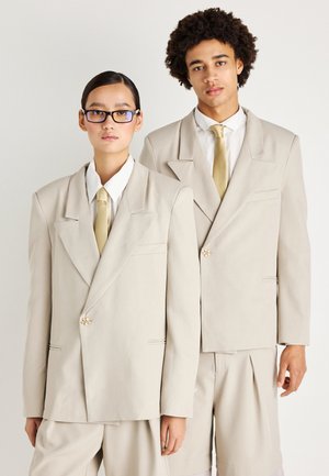 Dos modelos visten trajes oversized en color beige con solapas afiladas, botones frontales y corbatas amarillas pálidas a juego. La tela tiene una textura suave.