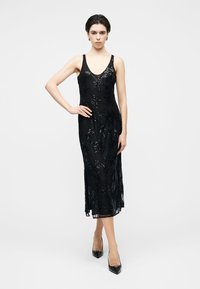 Μαύρο φόρεμα από δαντέλα με λεπτή εφαρμογή, V-λαιμόκοψη και περίπλοκες διακοσμήσεις με λουλούδια· μήκος έως το γόνατο με ελαφρώς διαφανή υφή· συνδυασμένο με μυτερά τακούνια.