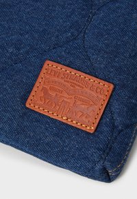 Tmavomodrá džínová látka s textúrovaným povrchom, s hnedou koženou nášivkou s embosovaným nápisom "Levi Strauss & Co." a dekoratívnym prešívaním.