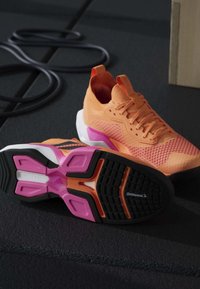 Paire de chaussures de sport orange avec des semelles roses et noires sur un sol sombre de salle de sport à côté d'une bande de résistance noire enroulée.