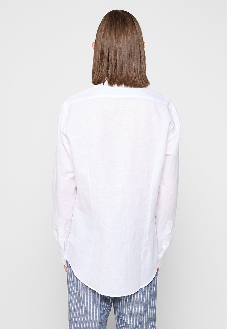 Biała lniana koszula z długim rękawem, z kołnierzykiem, o zaokrąglonym dolnym brzegu i luźnym kroju. Posiada subtelną teksturę oraz szew yoke z tyłu.