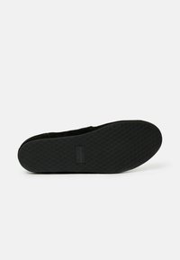 Scarpa nera slip-on con suola in gomma testurizzata che presenta un motivo a rombi e una piccola etichetta con logo sul lato. Il materiale sembra morbido e flessibile.