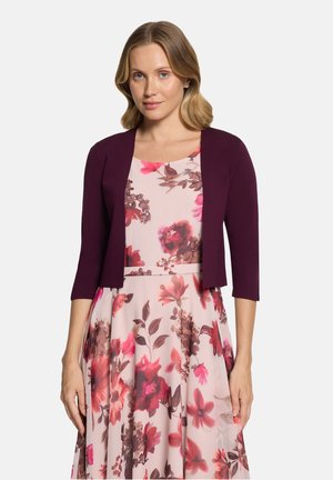 Kvinde med skulderlangt blondt hår iført en mørk lilla kort cardigan over en lyserød blomsterkjole med røde og brune blomster.
