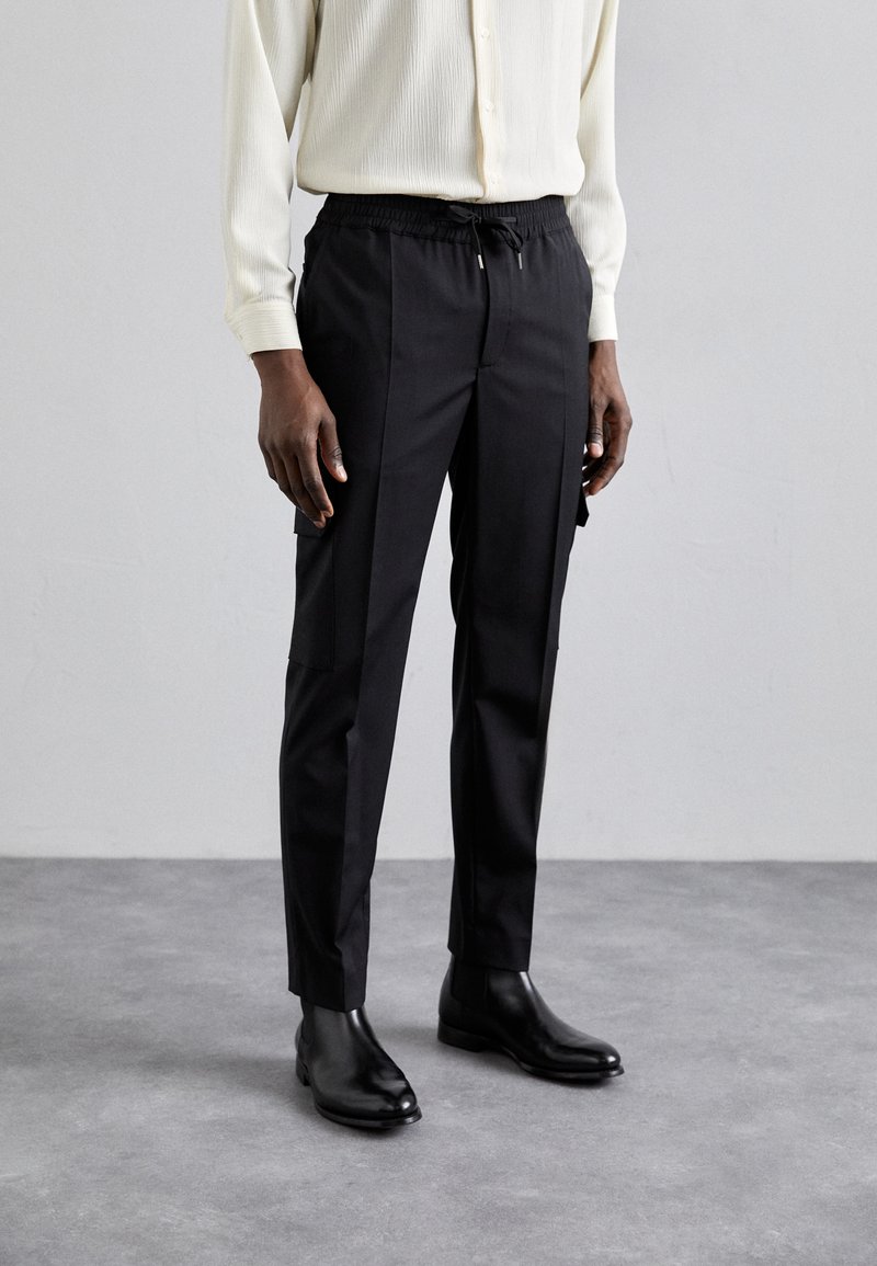 Zwarte tapered broek met een elastische tailleband en trekkoorden, voorzien van zijzakken en een soepele stofstructuur, gecombineerd met zwarte enkellaarzen.