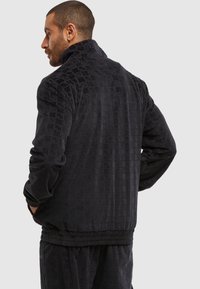 Homme avec les cheveux courts et une boucle d'oreille, portant une veste noire texturée et un pantalon assorti, les mains dans les poches, de profil contre un fond uni.