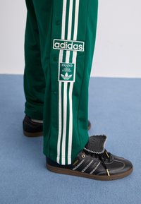 Grønne træningsbukser med hvide tre-stribs detaljer, Adidas-logoer og trykknaplukninger. Kombineret med sorte Samba-sneakers og teksturerede accenter.