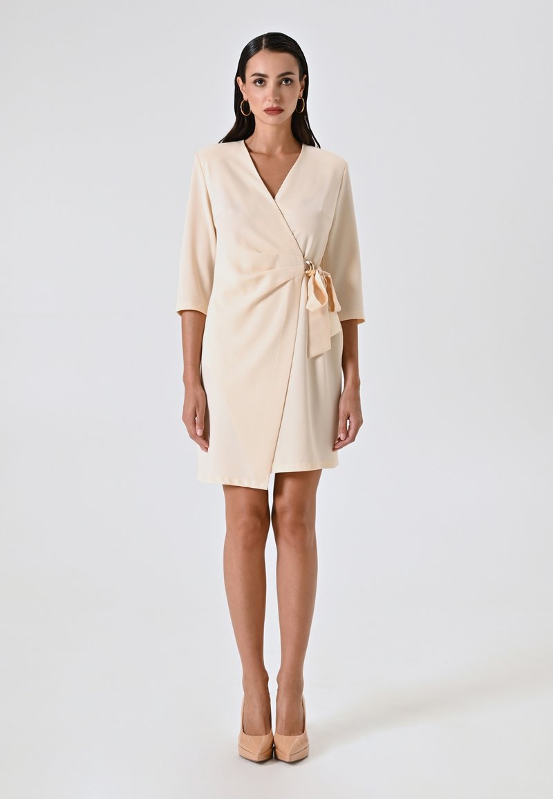 Beige Wickelkleid mit Dreiviertelärmeln, in der Taille mit einer weichen Schleife gebunden. Aus glattetem Stoff gefertigt, hat es einen V-Ausschnitt und eine Länge über dem Knie.