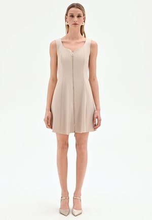 Femme portant une robe beige sans manches avec une fermeture éclair devant et des talons pointus métalliques, debout devant un fond blanc uni.