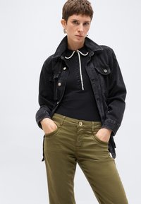 Γυναίκα με κοντά μαλλιά που φορά χρυσούς κρίκους στα αυτιά, μαύρο τζιν μπουφάν, μαύρο ριμπ τοπ με διακόσμηση από πέρλες και ελιά πράσινο παντελόνι με τα χέρια στις τσέπες.