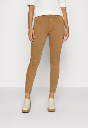 Schmal geschnittene braune Hose getragen mit beigen Plateau-Schuhen und einem weißen Langarmoberteil, gezeigt vom mittleren Oberkörper bis zu den Füßen vor einfachem Hintergrund.